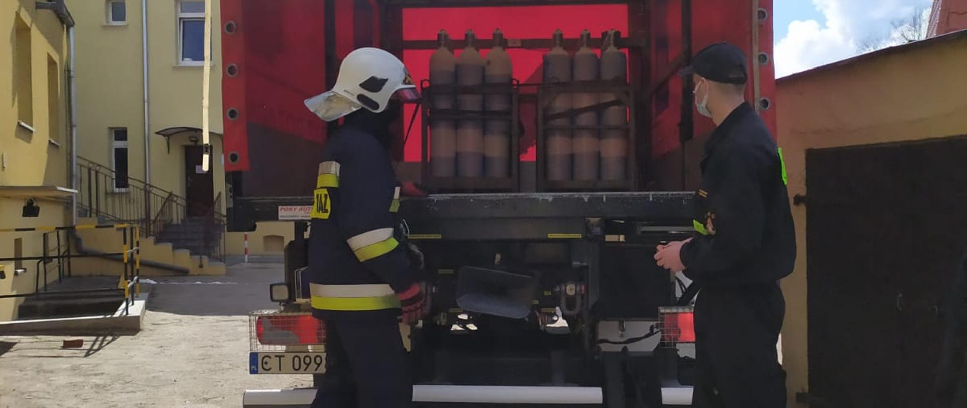 Na zdjęciu widoczny od tyłu czerwono-biały ciężarowy samochód strażacki stojący na utwardzonym terenie. Za pojazdem stoją dwaj strażacy. Jeden w mundurze koszarowym, drugi w mundurze bojowym i hełmie. Na otwartym przedziale ładunkowym znajdują się 2 metalowe kosze zawierające pakiet białych butli stalowych. W tle błękitne, częściowo zachmurzone niebo oraz budynki w kolorze kremowym.