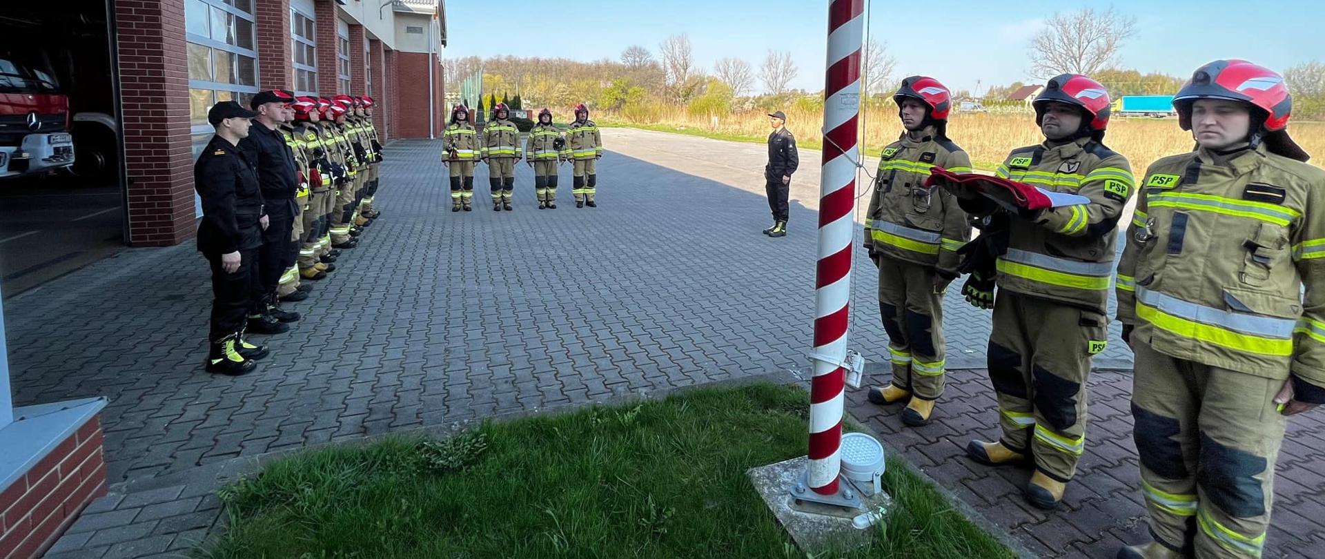 Na zdjęciu znajduje się uroczysta zmiana służby w której to strażacy ubrani w ubrania specjalne dokonują podniesienia flagi państwowej w obecności kierownictwa Komendy.