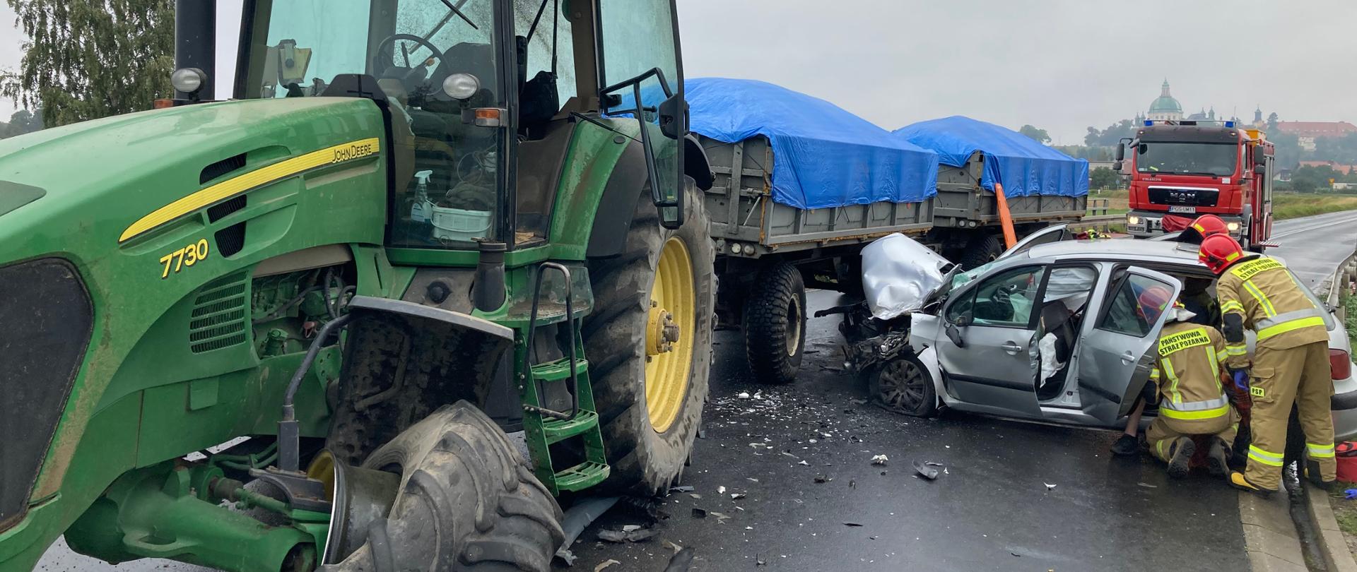 Zdjęcie przedstawia samochód osobowy, który uderzył w ciągnik rolniczy. Strażacy udzielają pomocy osobom poszkodowanym.