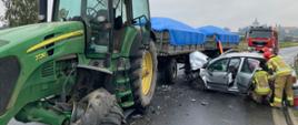 Zdjęcie przedstawia samochód osobowy, który uderzył w ciągnik rolniczy. Strażacy udzielają pomocy osobom poszkodowanym.