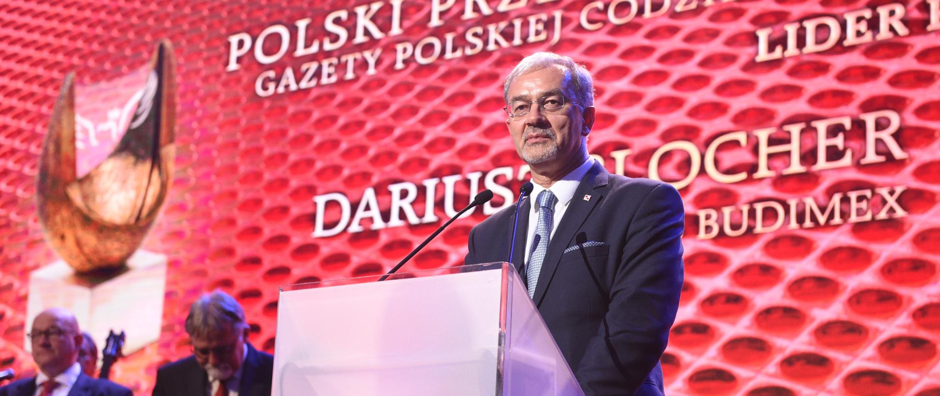 Na tle czerwonego ekranu z białym napisem "Polski Przedsiębiorca Gazety Polskiej Codziennie" stoi w mównicy minister Jerzy Kwieciński