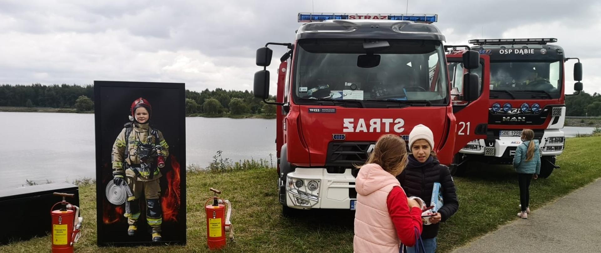 Zdjęcie przedstawia samochody ratowniczo- gaśnicze zaparkowane przy akwenie wodnym oraz dzieci które wykonywały zdjęcia przy makiecie strażaka.