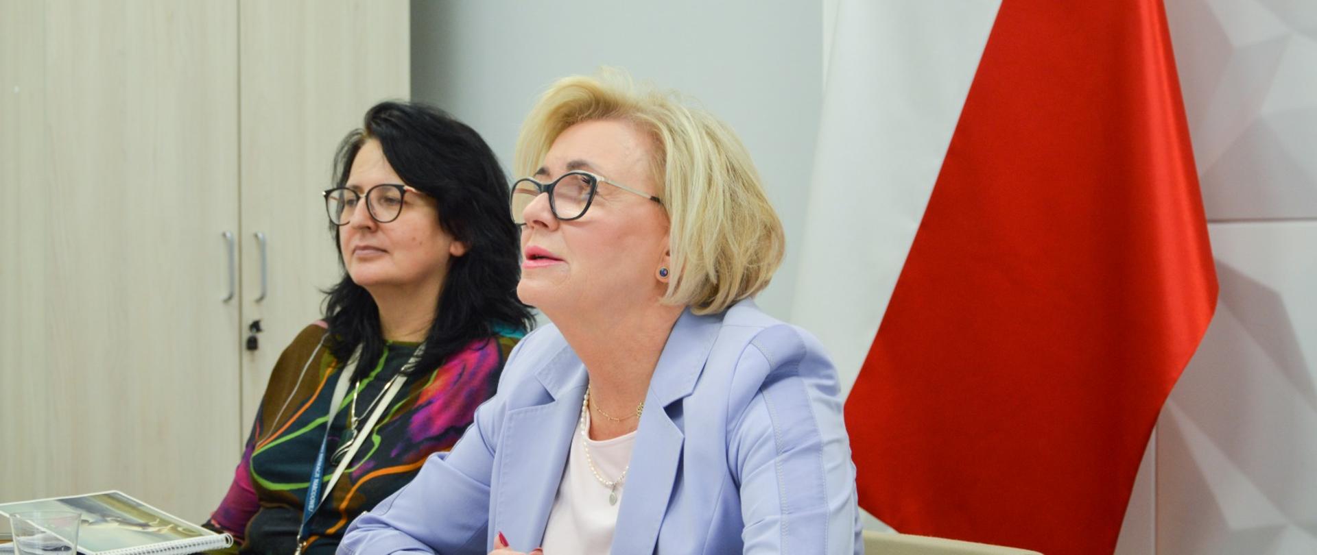 Wiceminister Machałek i kobieta z czarnymi włosami siedzą przy biurku, za nimi przy ścianie polska flaga.