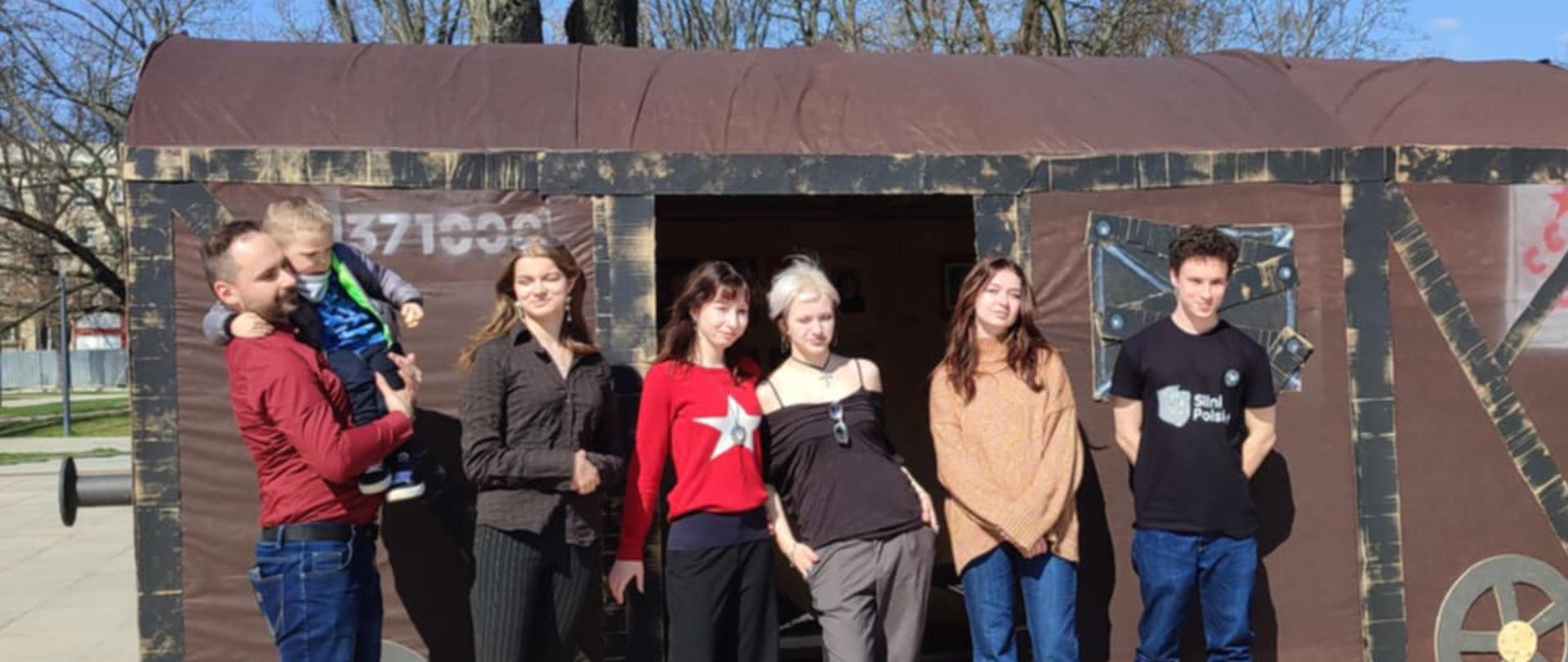 Na zdjęciu grupa osób stoi na zewnątrz przed drewnianą konstrukcją przypominającą historyczny wagon towarowy. Konstrukcja ma ciemne ściany z imitacją metalowych wzmocnień i koła na dole. Na ścianie widoczny jest numer „371003”. W tle znajdują się bezlistne drzewa i jasne, bezchmurne niebo, sugerujące wczesną wiosnę lub późną jesień.
Po lewej stronie nauczyciel Tomasz Banaszkiewicz w czerwonej bluzie trzyma na rękach małe dziecko ubrane w niebieską kurtkę i zielony plecaczek. Obok niego stoi uczennica w ciemnym stroju. W centrum znajdują się trzy uczennice – jedna w czerwonym swetrze z białą gwiazdą, druga w czarnym stroju na ramiączkach z okularami zawieszonymi na szyi, a trzecia w luźnym brązowym swetrze i dżinsach. Po prawej stronie stoi uczeń ubrany w czarną koszulkę z napisem „Sinti Polska” oraz granatowe spodnie.
Wszyscy stoją blisko siebie i patrzą w stronę aparatu, a ich postawa sugeruje swobodę i uczestnictwo w wydarzeniu o edukacyjnym lub historycznym charakterze.