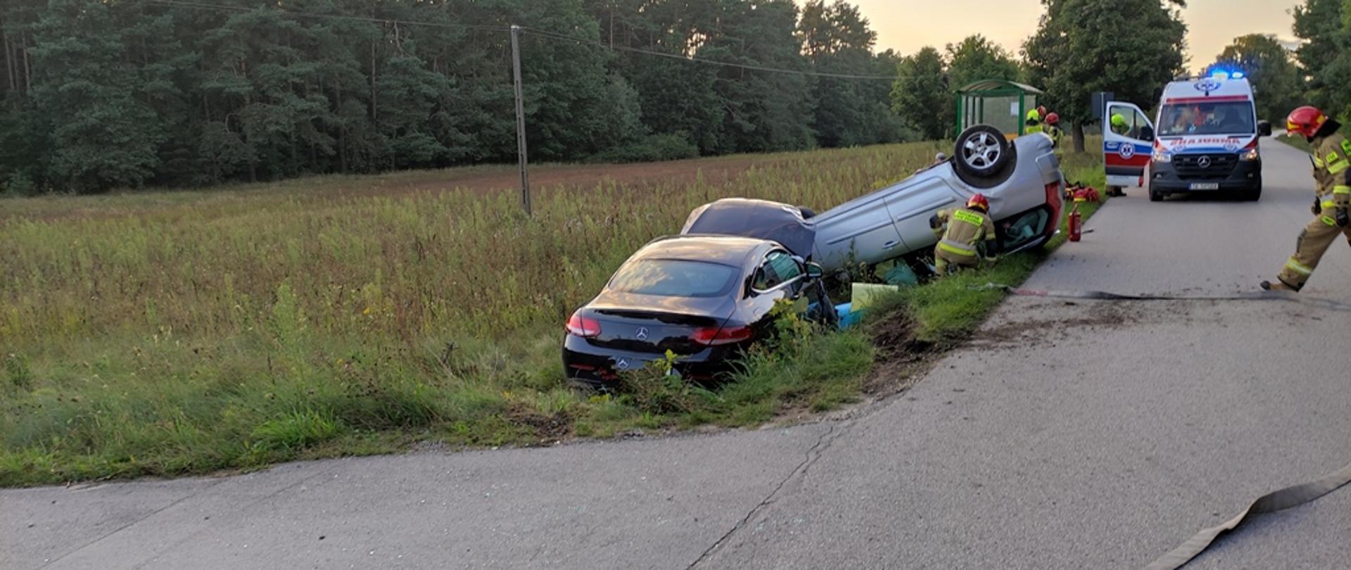 Droga powiatowa, zderzenie dwóch samochodów osobowych, pojazdy znajdują się w przydrożnym rowie, Mercedes na kołach, a Citroen na dachu, dwóch strażaków w ubraniach specjalnych dokonuje sprawdzenia pojazdów. Na drugim planie karetka pogotowia oraz przystanek autobusowy.