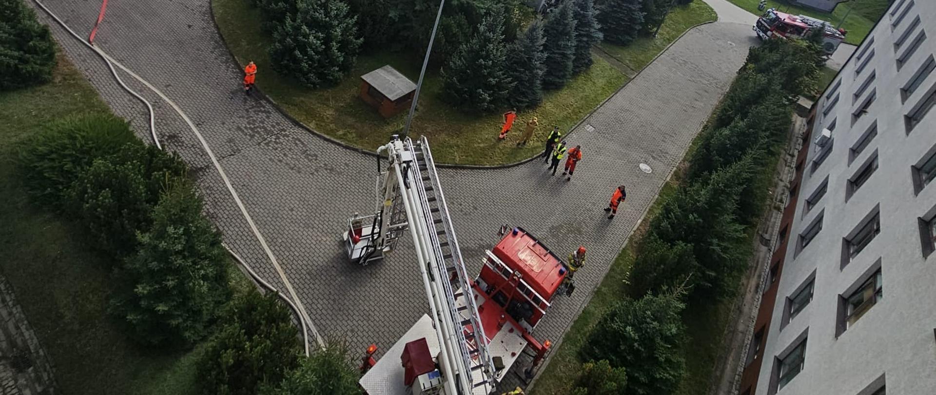 Zdjęcie przedstawia widok z góry na plac przed szpitalem, w centrum widać samochód specjalny podnośnik z drabiną, oraz ćwiczących strażaków po prawej i po lewej stronie widoczne samochody strażackie.