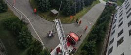 Zdjęcie przedstawia widok z góry na plac przed szpitalem, w centrum widać samochód specjalny podnośnik z drabiną, oraz ćwiczących strażaków po prawej i po lewej stronie widoczne samochody strażackie.