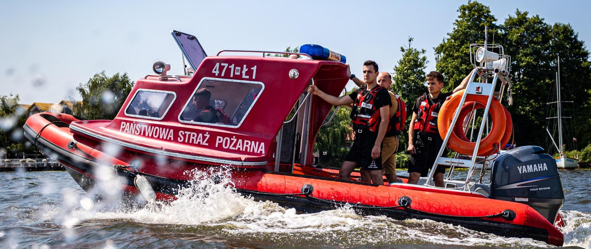 Na łodzi czterech strażaków - jeden w kajucie przy sterze, trzech stoi ma pokładzie. Łódź czerwona, z napisem Państwowa Straż Pożarna. Płyną po akwenie, w tle drzewa...
