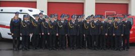 Wszyscy strażacy biorący udział w uroczystości, w tle samochody busy Komendy i jasna elewacja budynku
