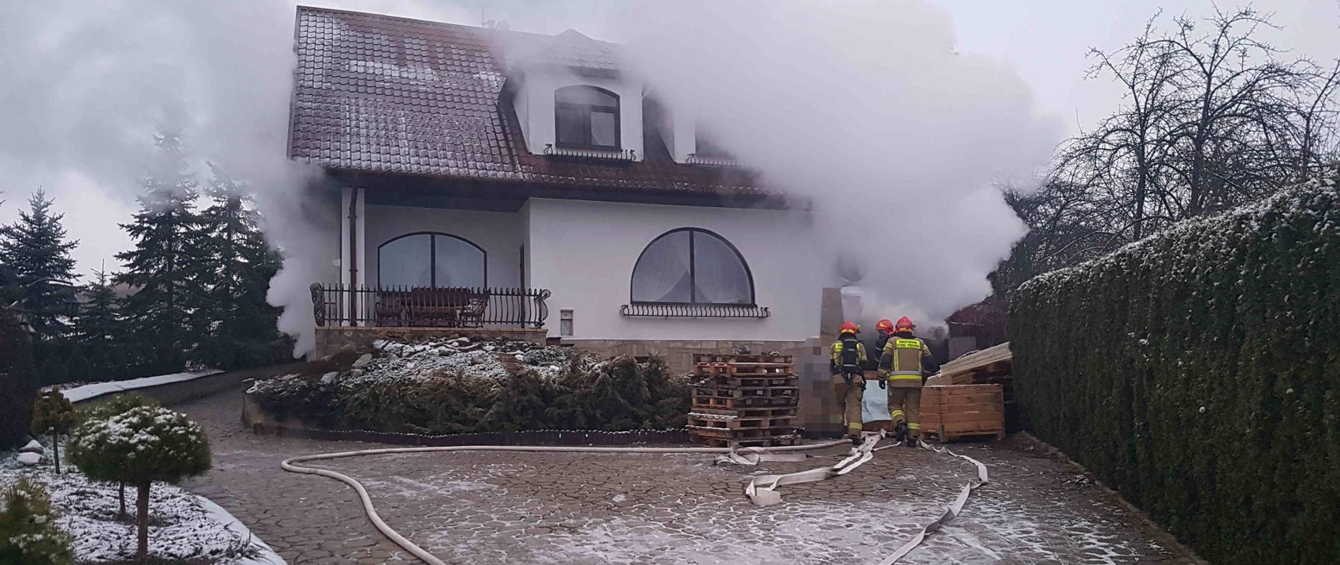 Zdjęcie zrobione w dzień. Na zdjęciu widać dom jednorodzinny, z kotłowni wydobywa się dym. Przed domem stoją strażacy z nawodnioną linią gaśniczą, którzy są przygotowani do wejścia do kotłowni w celu ugaszenia pożaru.