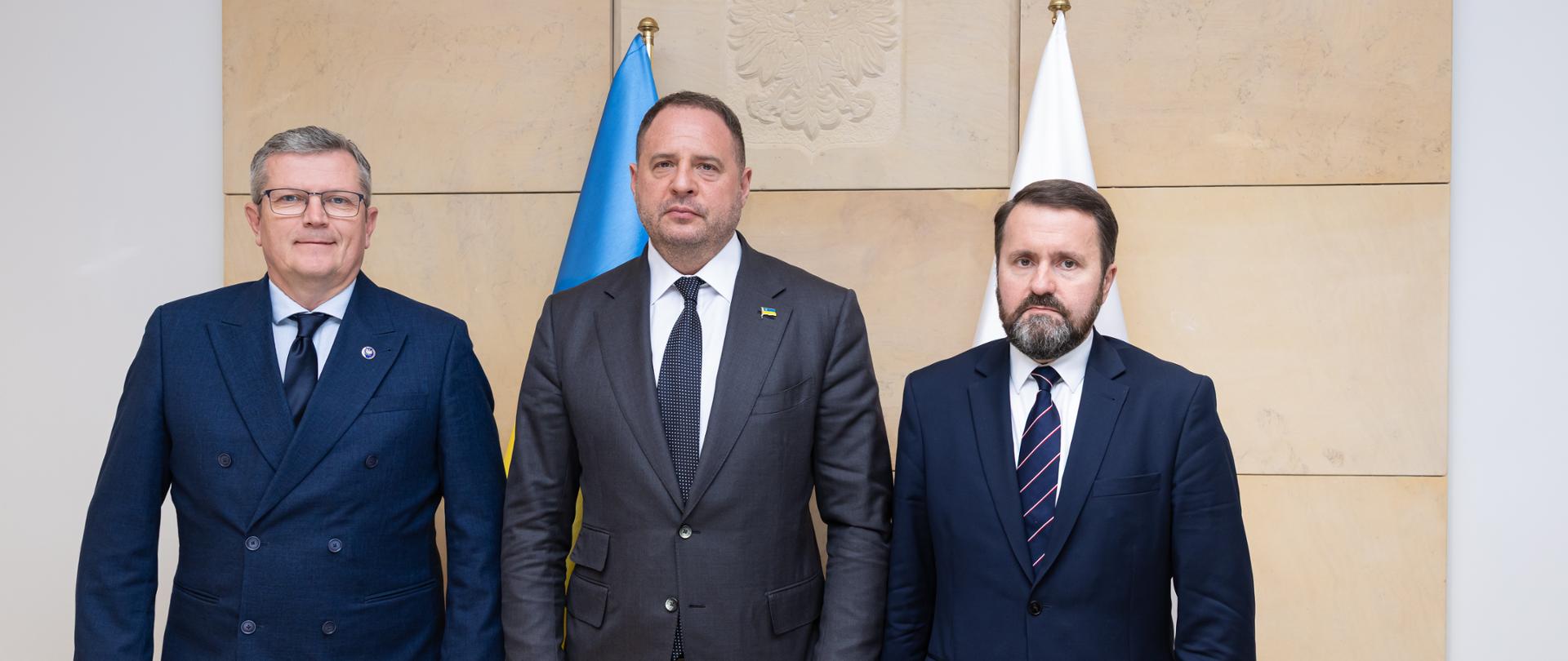 Na zdjęciu jest trzech mężczyzn ubranych w ciemne garnitury. Stoją na jasnym tle. Za nimi są dwie flagi - Polski i Ukrainy. 