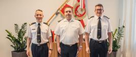Gabinet Komendanta. Na pierwszym planie ubrani w mundury galowe tj. białe koszule i czarne spodnie stoją: Komendant Główny nadbrygadier Andrzej Bartkowiak, nowo powołany zastępca dyrektora w Biurze Finansów w Komendzie Głównej Państwowej Straży Pożarnej brygadier Jarosław Kociołek, Zastępca Komendanta Głównego PSP nadbrygadier Krzysztof Hejduk. W tle sztandar PSP. 