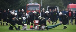 Zdjęcie przedstawia 6 druhów OSP podczas ćwiczeń bojowych na zawodach sportowo -pożarniczych. Dwóch z nich trzyma węże ssawne, pozostali chwytają w dłonie węże pożarnicze z drewnianej platformy. Ubrani są w stroje koszarowe i białe hełmy. Wszystko dzieje się na boisku do piłki nożnej. W oddali widać parking z samochodami strażackimi oraz innych druhów OSP.