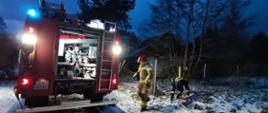 Na zdjęciu dwóch strażaków. Jeden z nich pochylony łączy odcinki węża na ośnieżonej drodze, drugi zbliża się do widocznego po lewej stronie zdjęcia stojącego i oświetlonego sygnałami alarmowymi tyłu wozu pożarniczego z widoczną autopompą. W tle budynek mieszkalny o zmierzchu.