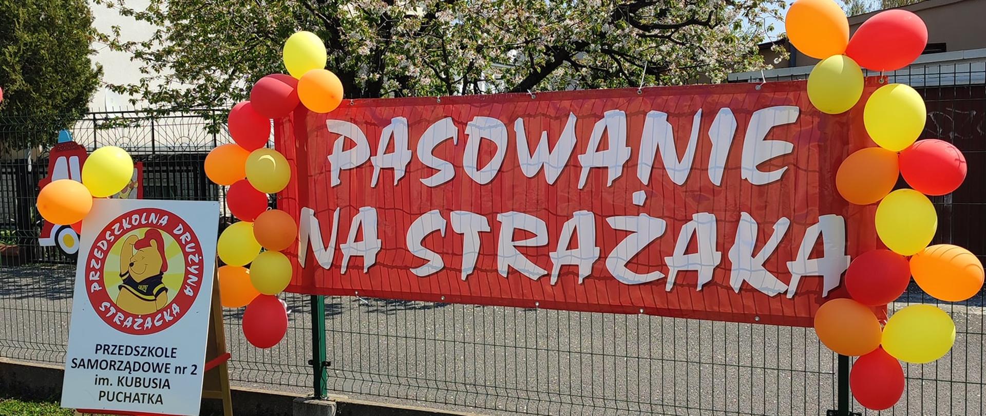 Zdjęcie przedstawia na pierwszym planie czerwoną szarfę z napisem Pasowanie na strażaka. Dookoła szarfy umieszczono czerwono żółte balony. Dookoła tereny zielone.