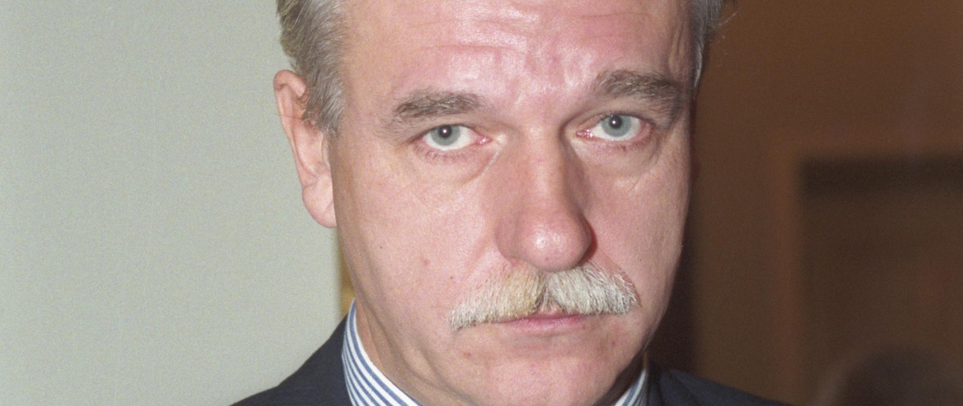 Andrzej Olechowski kolorowy portret mężczyzny, siwe włosy, wąsy, granatowa marynarka w paski, granatowy krawat w czerwone paski, biała koszula w granatowe paski.