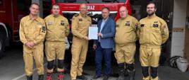 Druhowie OSP Gorce, Komendant Miejski PSP w Wałbrzychu oraz Z-ca Burmistrza Miasta Boguszów-Gorce pozują do zdjęcia na tle czerwonego wozu strażackiego stojącego w remizie.