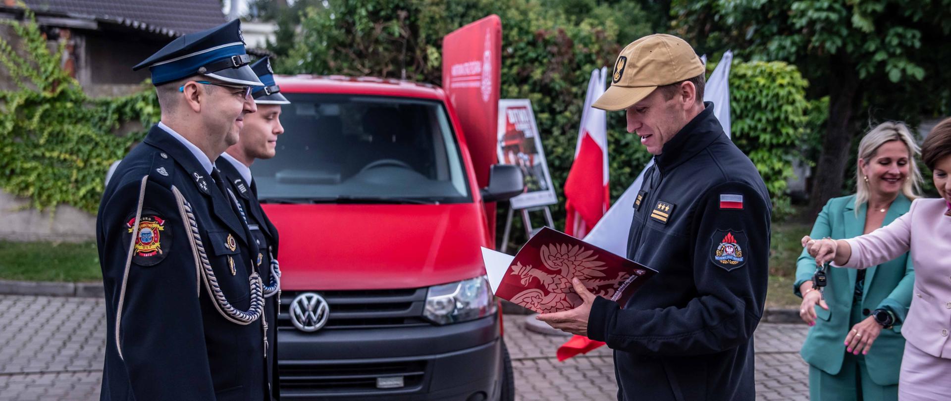 strażak odczytuje list gratulacyjny , przed nim stoi dwóch druhów OSP, w tle czerwony samochód strażacki