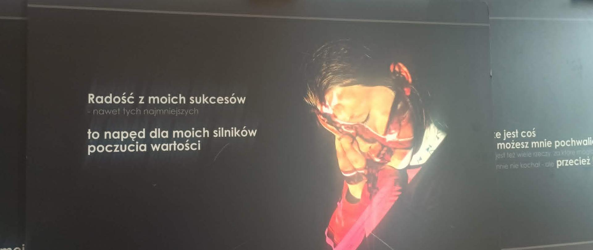 Zbliżenie na czarną planszę wystawową z fotografią osoby zakrywającej twarz dłońmi. Obok zdjęcia widoczny jest polski cytat zaczynający się od słów ‘Radość z moich sukcesów…’, odnoszący się do poczucia własnej wartości. Plansza stoi na drewnianej sztaludze.