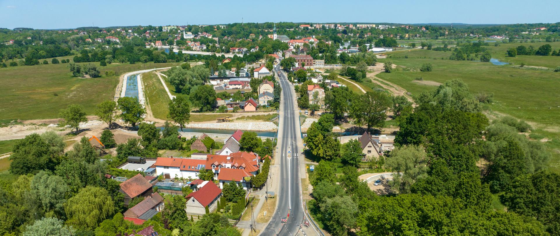 Droga krajowa jednojezdniowa w otoczeniu zabudowy domów jednorodzinnych i zieleni.