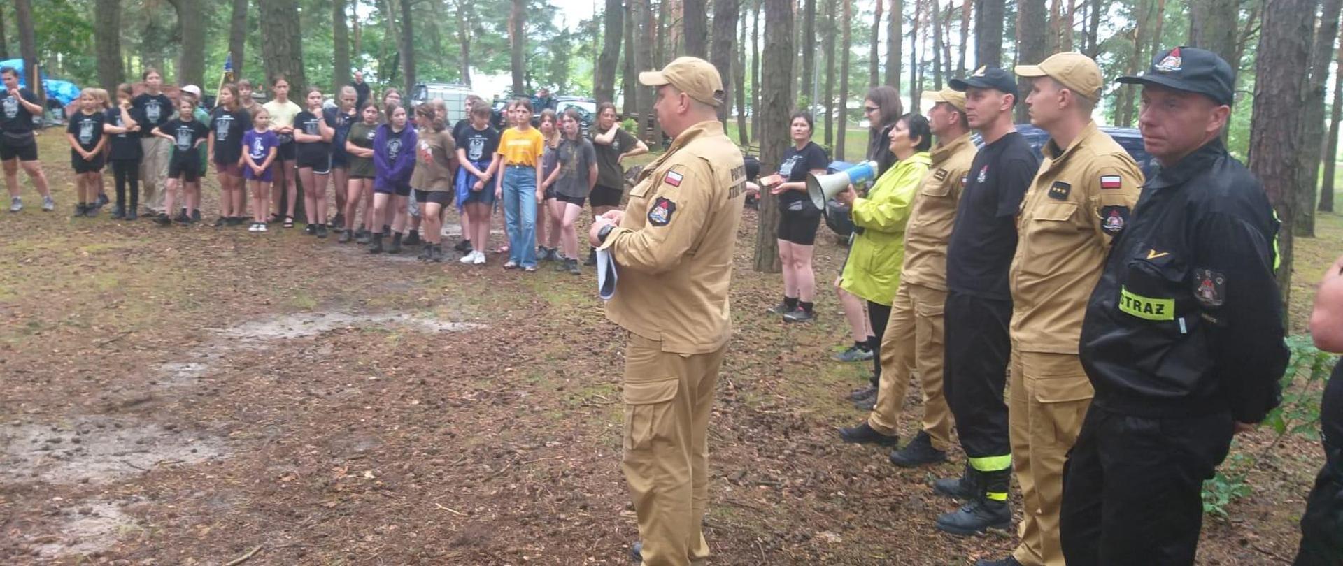 Zdjęcie przedstawia spotkanie strażaków z wypoczywającą młodzieżą szkolną w lesie gdzie urządzony został obóz harcerski.