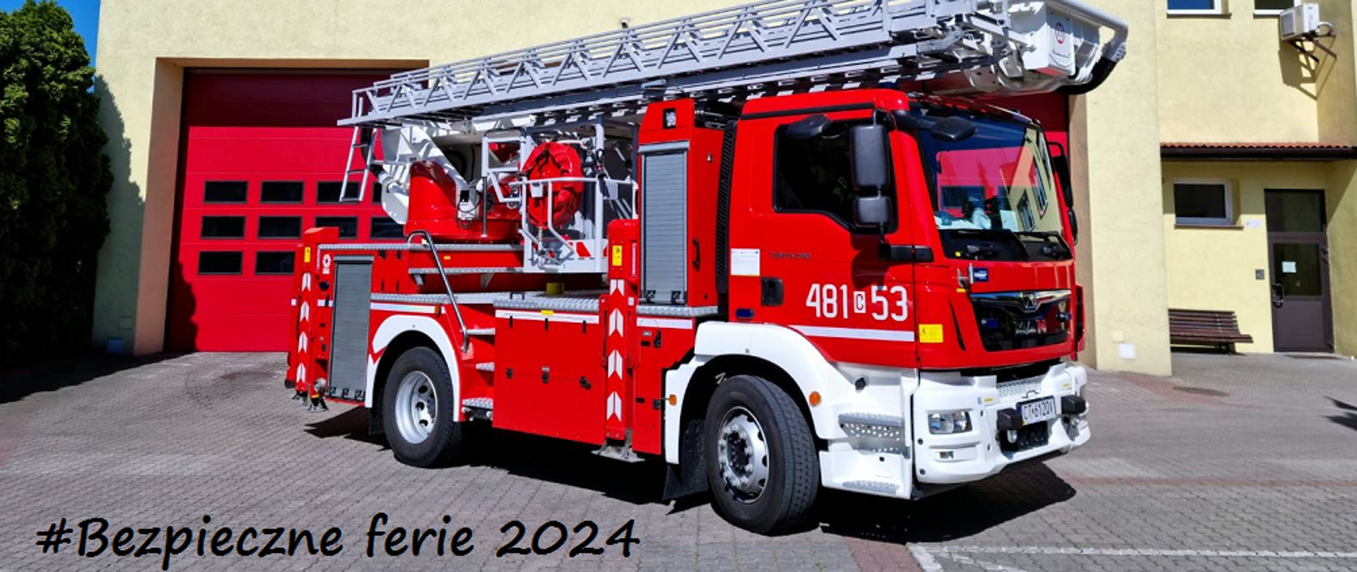 Samochód strażacki - podnośnik hydrauliczny na tle budynku komendy. Po lewej stronie napis "Bezpieczne ferie 2024"