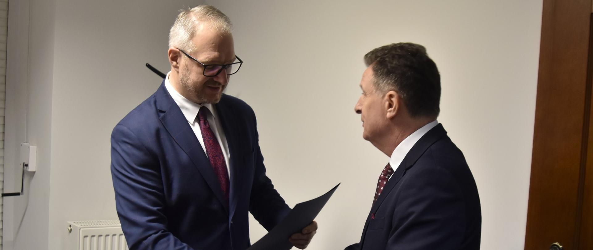 zdjęcie przedstawia dwóch mężczyzn w garniturach. Jeden z nich przekazuje granatowa teczkę drugiemu mężczyźnie. 