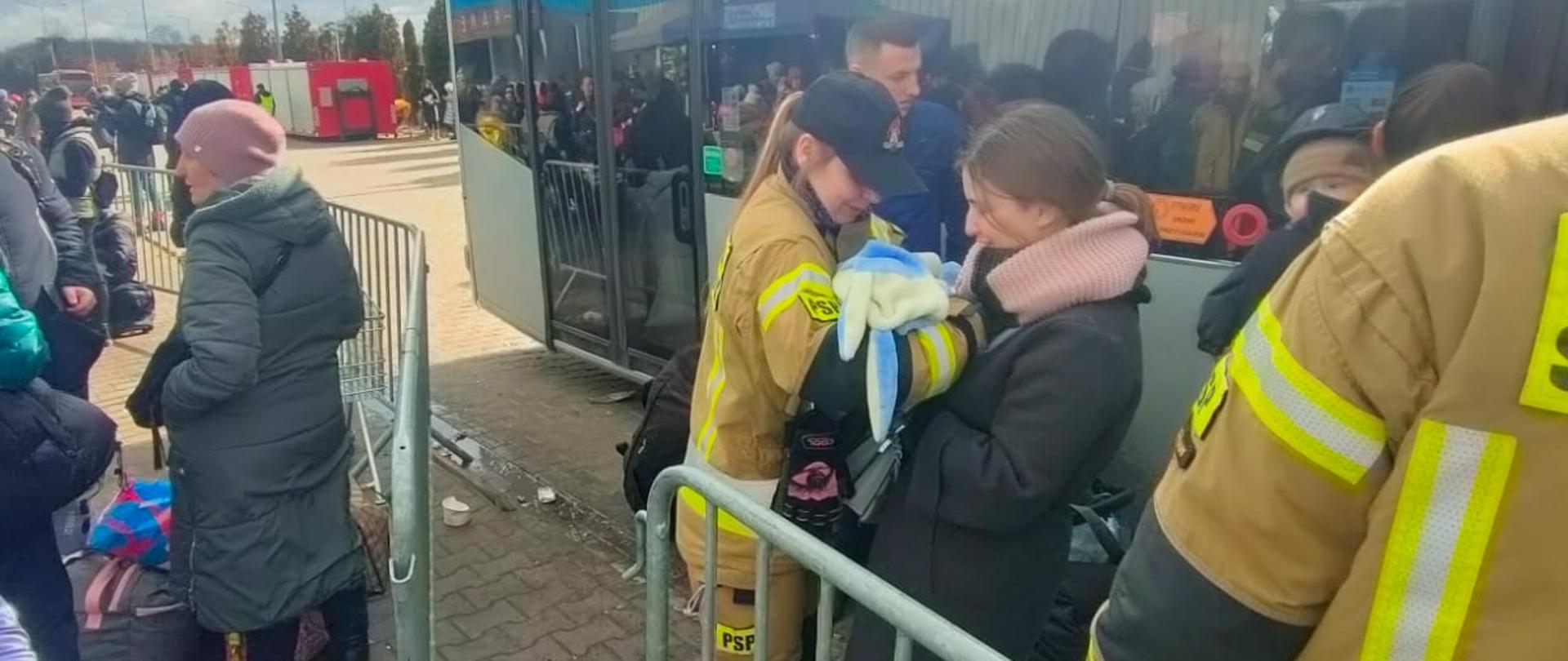 Na zdjęciu widać kobietę, która przekazuje strażakowi niemowlę. Dookoła widać innych uchodźców, a także strażaków. Z tyłu stoi zaparkowany autobus.