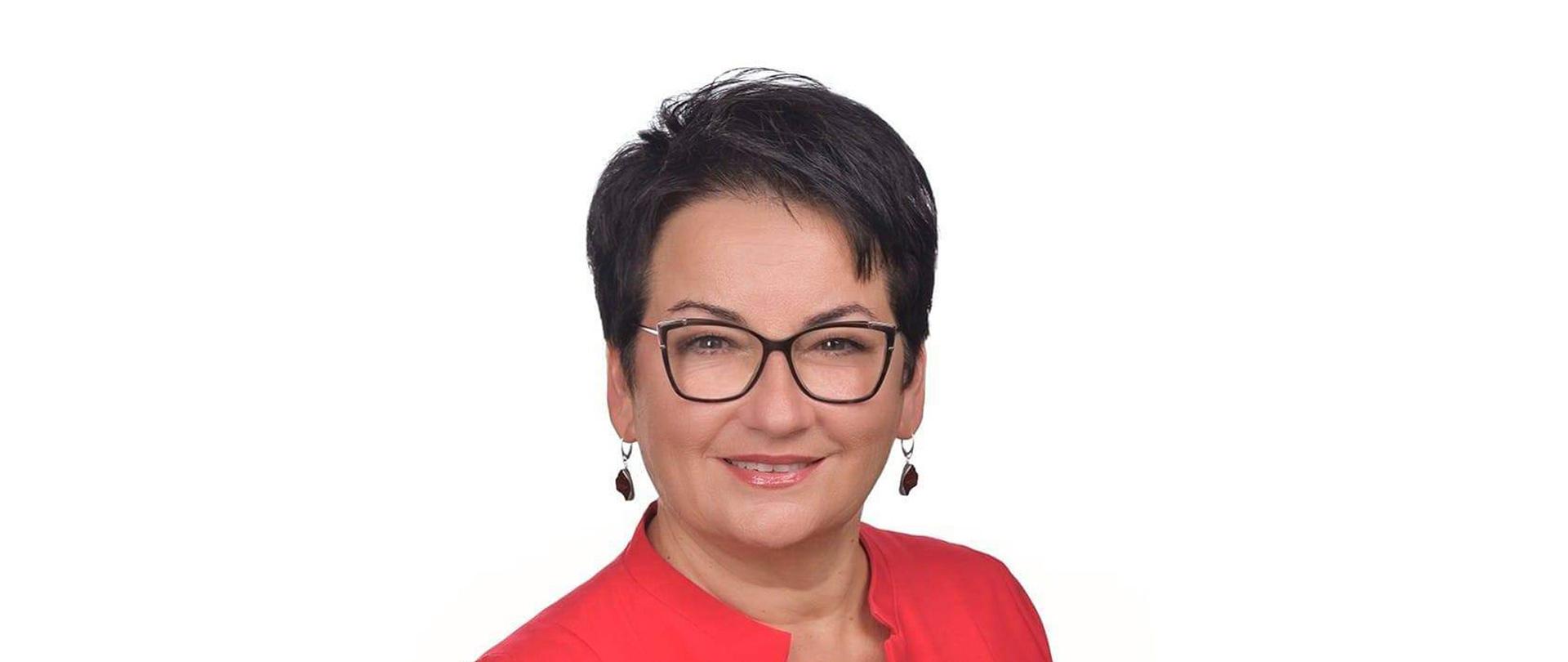 Portret uśmiechnięty kobiety. Ma krótkie ciemne włosy. Ubrana jest w czerwona bluzkę. Na nosie ma czarne okulary korekcyjne.