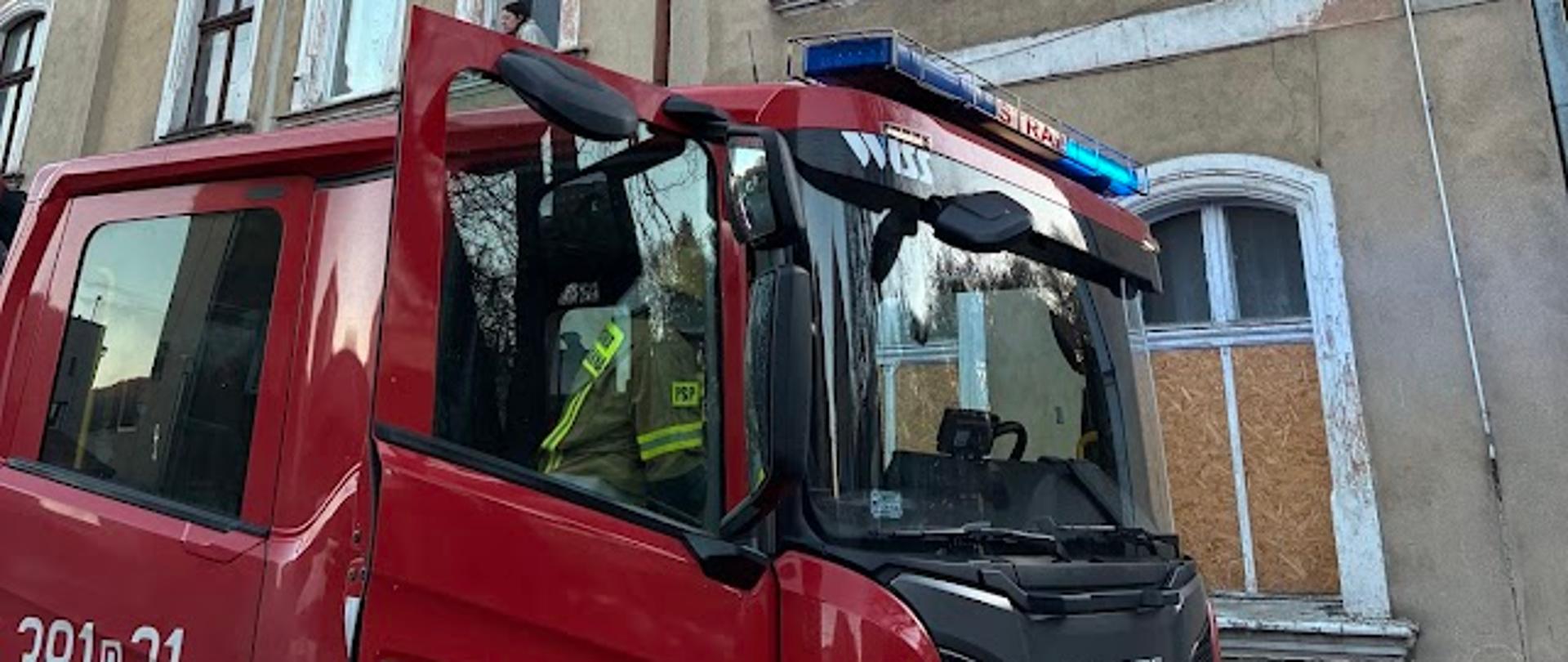 Zdjęcie przedstawia czerwony wóz strażacki marki Scania zaparkowany na wąskiej ulicy przed starą, wielopiętrową kamienicą o szarej elewacji. Pojazd ma włączone niebieskie sygnały świetlne, a w oknie kabiny widać strażaka w żółtym stroju ochronnym. Kamienica ma widoczne ślady upływu czasu – łuszczącą się farbę i zdobienia w stylu muru pruskiego na najwyższym piętrze. Jedno z okien na parterze jest zabite płytą OSB, co sugeruje wcześniejsze uszkodzenia lub zabezpieczenie miejsca zdarzenia