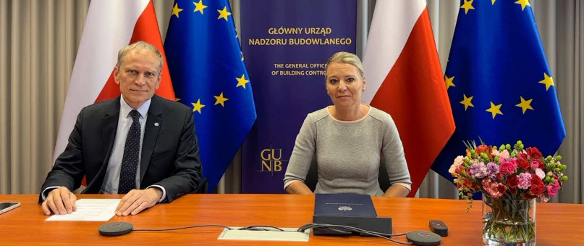 „Dwie osoby siedzące przy stole z mikrofonami i dokumentami, w tle baner GUNB oraz flagi Polski i UE, na stole bukiet kwiatów.”