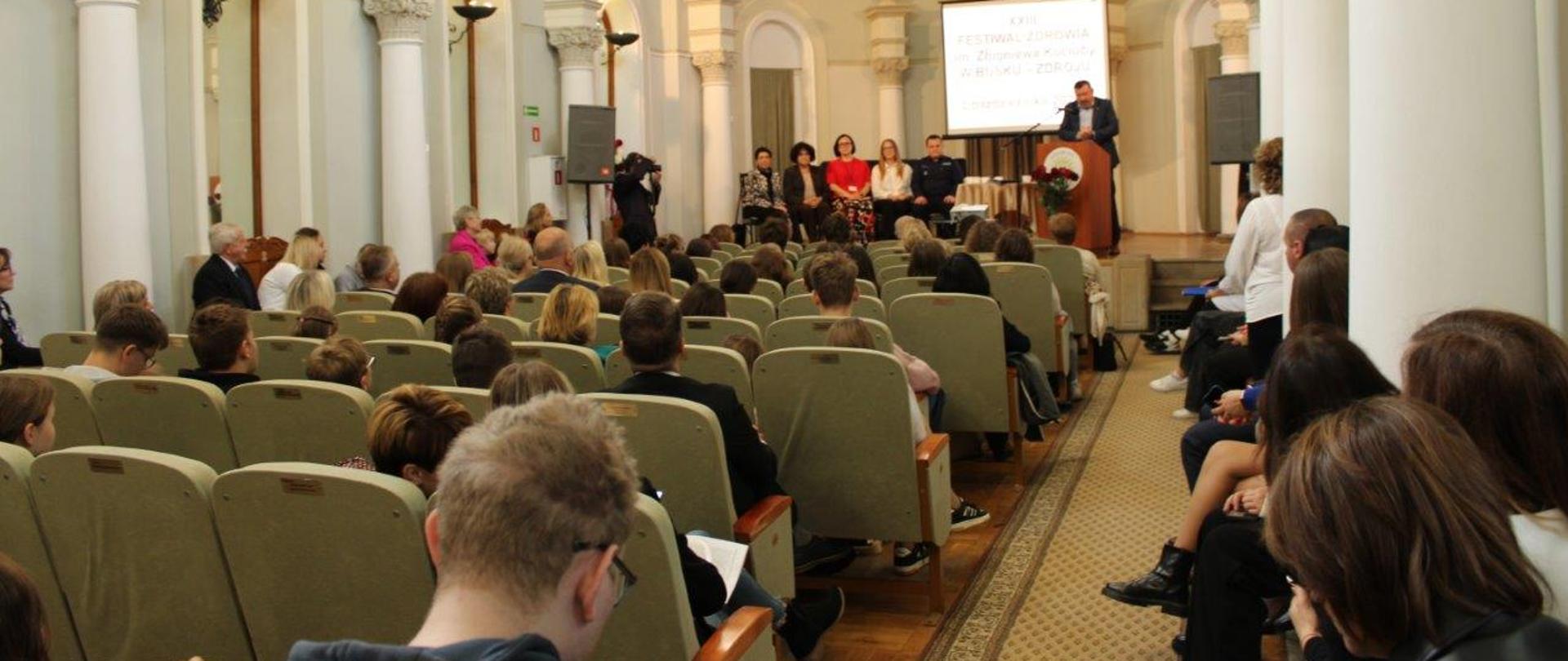 Na pierwszym planie słuchacze panelu dyskusyjnego "Jak zdrowo jeść?". W tle przy mównicy słowo wstępu głosi Świętokrzyski Państwowy Wojewódzki Inspektor Sanitarny a na scenie siedzą przedstawiciele różnych instytucji zajmujących się zdrowiem i bezpieczeństwem.