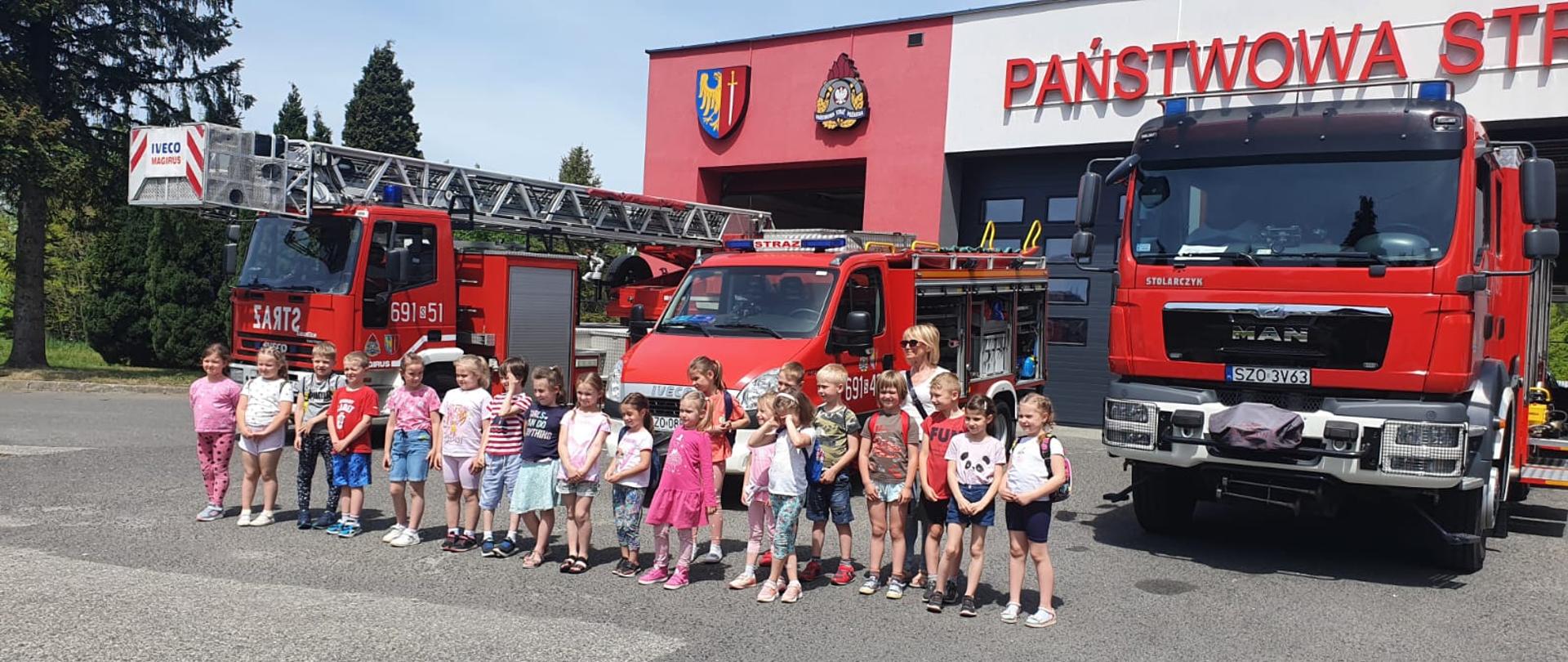 Dzieci i strażacy stoją przy samochodach strażackich. Strażacy opowiadają i pokazują sprzęt. 