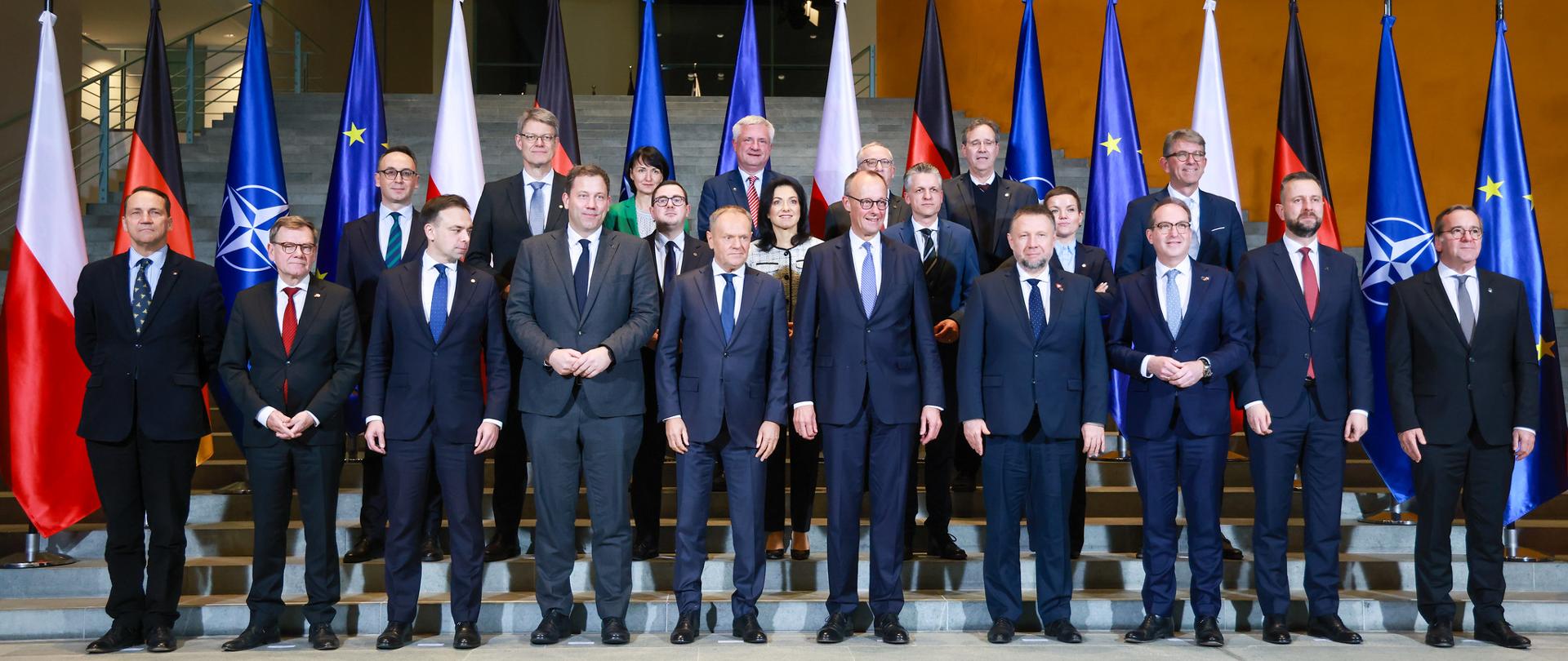 22 osoby w formalnych ubraniach pozujące do zdjęcia grupowego. W tle flagi PL, DE, NATO i UE.