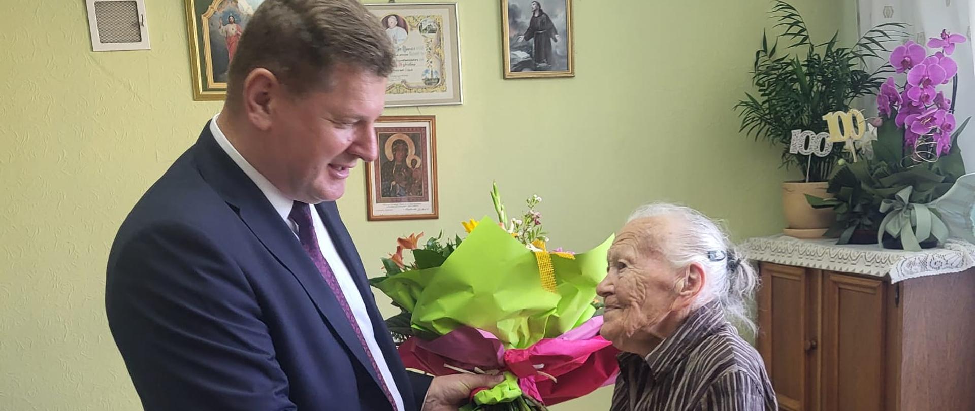 W pomieszczeniu znajdują się dwie osoby. Od lewej: Kierownik PT KRUS w Jarosławiu trzyma ręce trzyma bukiet kwiatów i wręcza je Pani Marii.