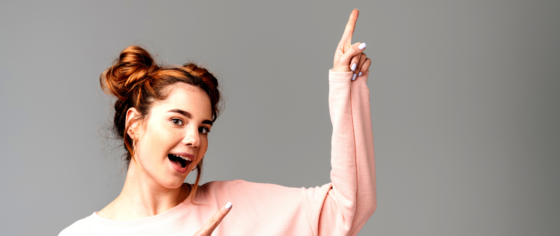 Uśmiechnięta ruda dziewczyna w pudrowej bluzie wskazująca rękoma w górę