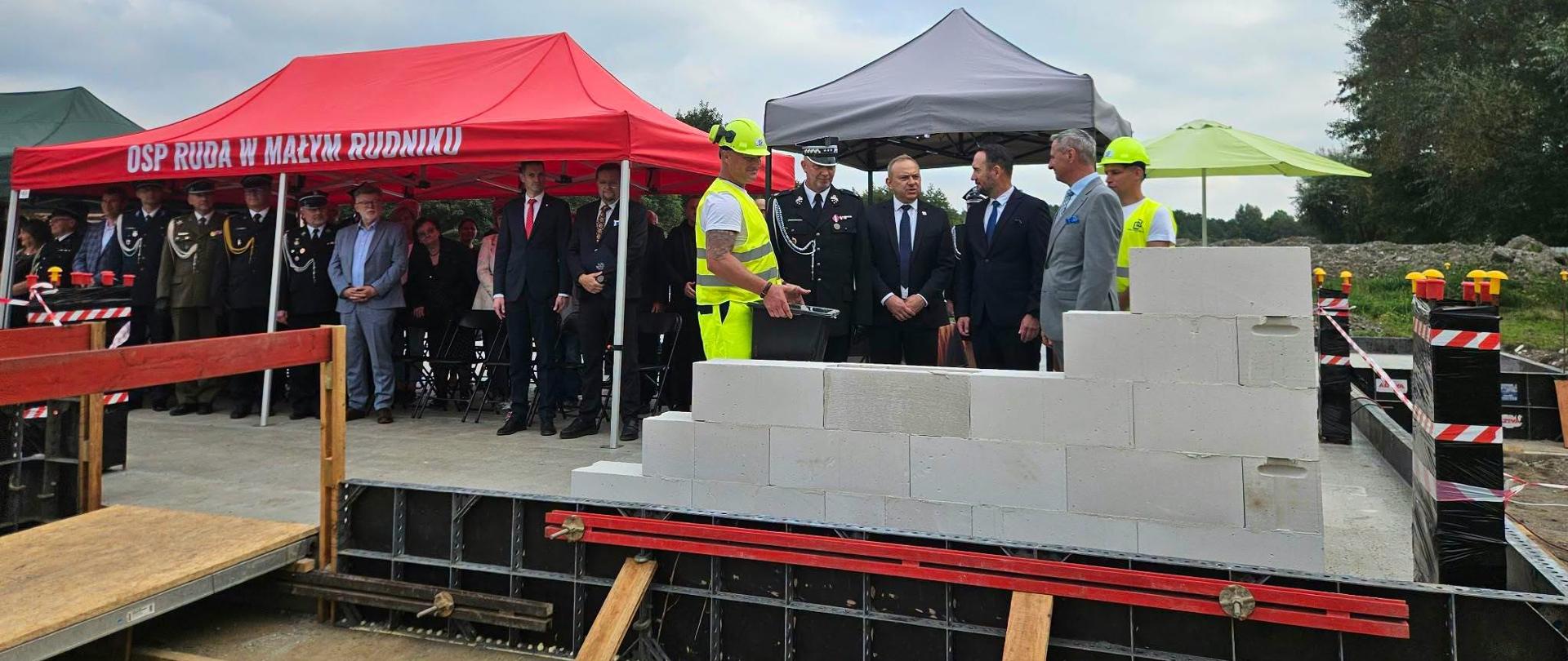 Zgromadzenie ludzi na terenie budowy. Przemowa oficjela przy rozpoczętej murowanej ścianie.