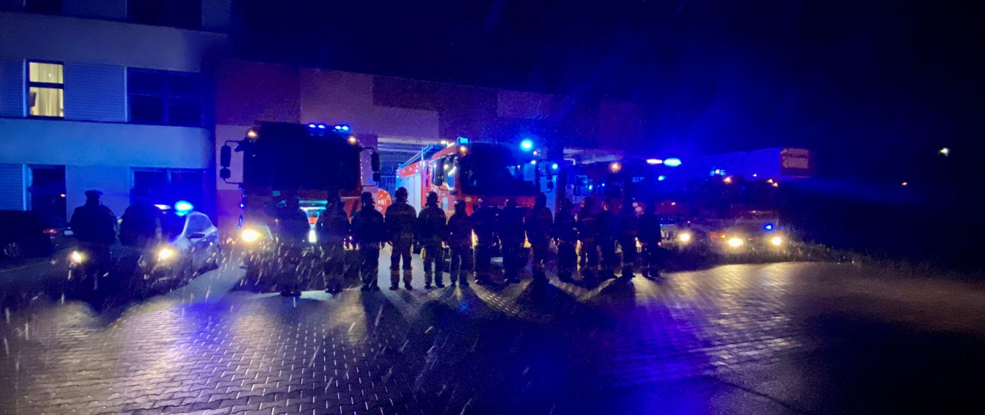 W strugach deszczu stoi zmiana służbowa JRG Stargard na tle samochodów pożarniczych z włączonymi światłami błyskowymi, po ich lewej stronie dwóch policjantów przed radiowozem również z włączonymi światłami błyskowymi. Na dalszym planie KP PSP Stargard.