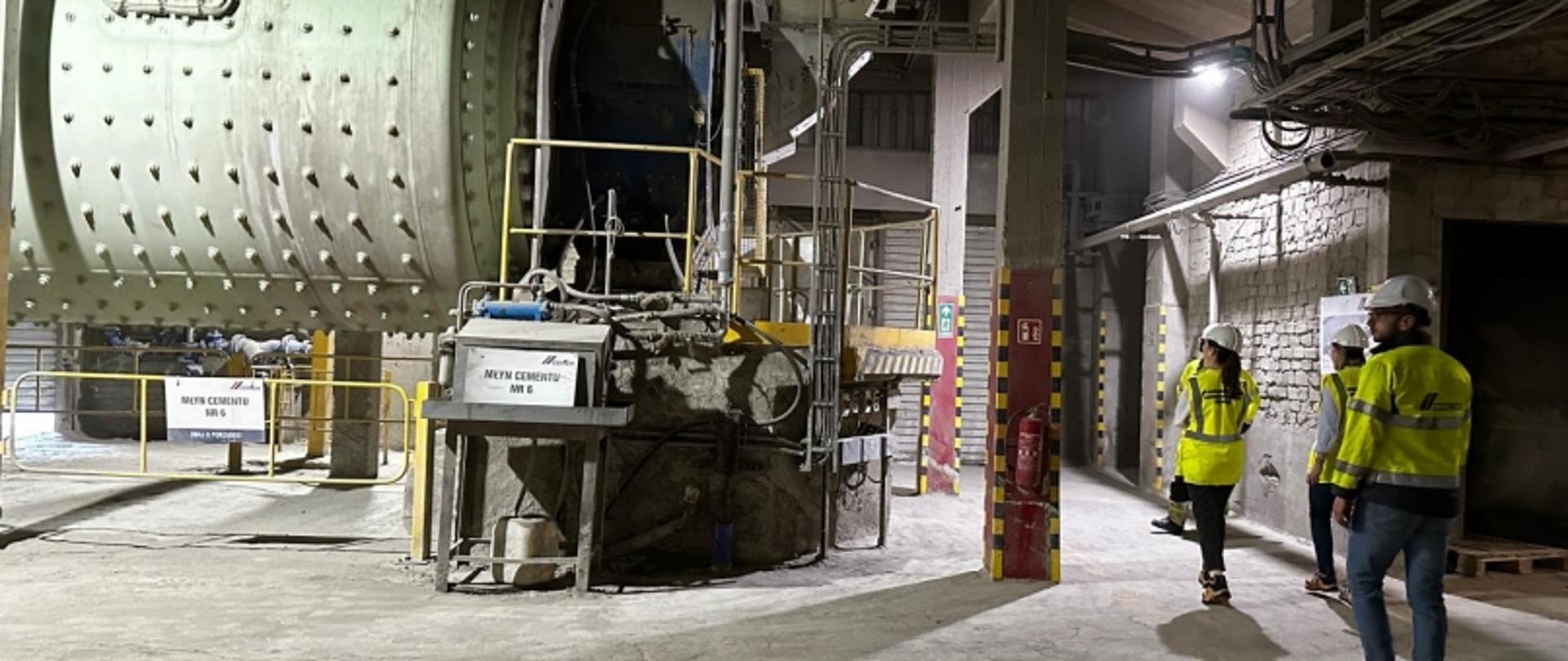 Uczestnicy wizyty technicznej zwiedzają cementownię – zarówno wnętrza zakładu, jak i infrastrukturę zewnętrzną. Ubrani w odzież ochronną, w tym kamizelki odblaskowe i kaski, zapoznają się z procesem produkcji cementu oraz biorą udział w prezentacji edukacyjnej.