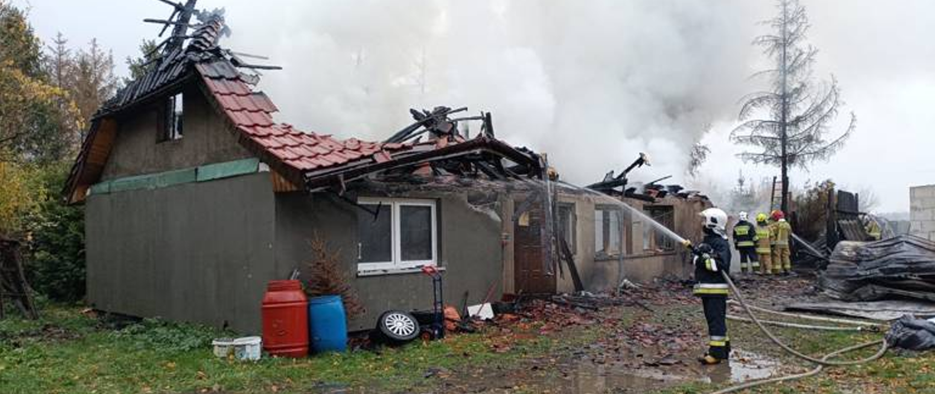 Strażacy gaszą ogień w częściowo zawalonym domu z zielonymi ścianami i czerwonym dachem. Widać wypalone fragmenty konstrukcji dachu i rozrzucone dachówki. Na ziemi leżą węże strażackie i pojemniki.