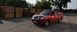 Zdjęcie przedstawia lekki samochód rozpoznawczo- ratowniczy marki Nissan Navara, który jako pierwszy dojeżdża używając sygnałów świetlnych do działań ratowniczych. W tle złożone palety z deskami.