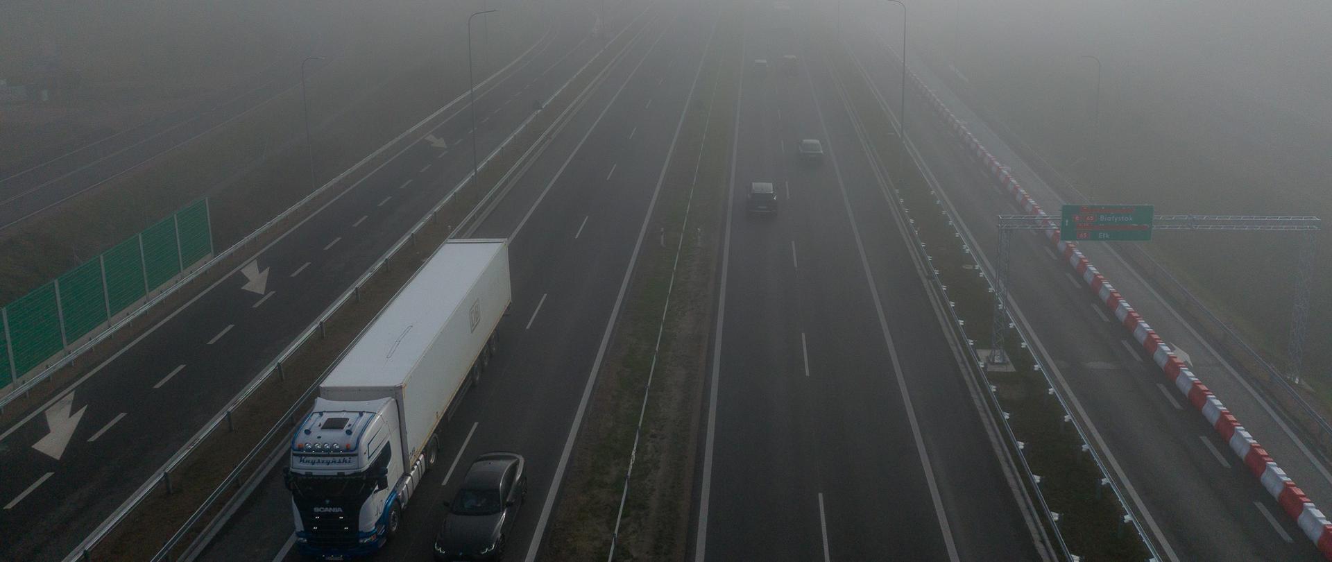Widok z góry na drogę ekspresową podczas gęstej mgły. Na jezdni poruszają się samochody osobowe i ciężarowe, a ograniczona widoczność utrudnia obserwację dalszego przebiegu trasy.