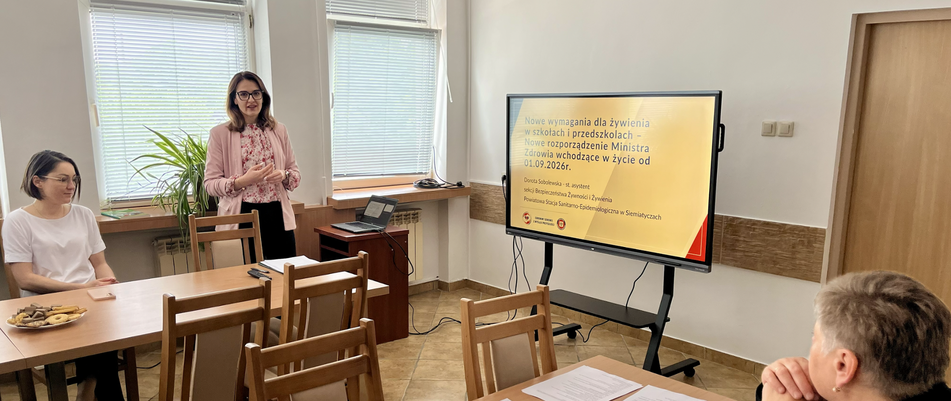 Pracownica Sekcji Bezpieczeństwa Żywności i Żywienia Powiatowej Stacji Sanitarno-Epidemiologicznej w Siemiatyczach prowadzi szkolenie dotyczące nowych przepisów żywieniowych dla szkół i przedszkoli. Obok prelegentki znajduje się monitor z prezentacją zatytułowaną „Nowe wymagania dla żywienia w szkołach i przedszkolach – nowe rozporządzenie Ministra Zdrowia wchodzące w życie od 01.09.2026 r.”. Uczestnicy szkolenia słuchają wykładu przy stołach z materiałami informacyjnymi.