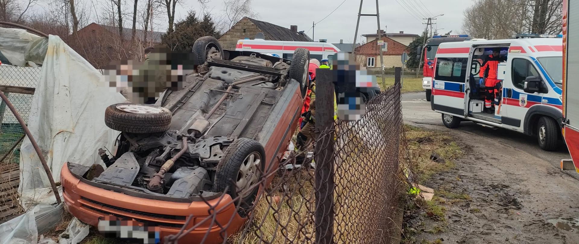 Na zdjęciu widać rozbity samochód osobowy, który leży na dachu za ogrodzeniem z siatki. Obok widać tunel foliowy. Na drodze stoją dwa samochody strażackie oraz dwie karetki pogotowia. W tle widać budynki, drzewa oraz latarnie. Jest widno, ale pochmurnie.