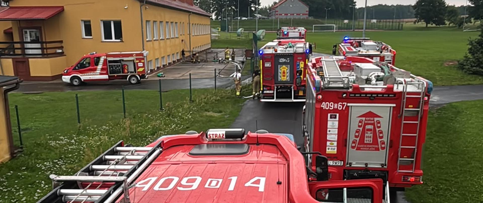 Spojrzenie z góry na akcję ratunkową prowadzoną przez strażaków. Z dachu wozu bojowego widać kilka pojazdów straży pożarnej (w tym dwa z wyraźnymi numerami operacyjnymi "409-14" i "409-67") zaparkowanych przed jednopiętrowym, żółtym budynkiem z czerwonym dachem. Budynek ten to prawdopodobnie szkoła lub inna placówka użyteczności publicznej. Na chodniku przy budynku widać węże strażackie i pojedynczych ratowników. W oddali rozpościera się widok na pole i drzewa pod zachmurzonym niebem.