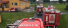 Spojrzenie z góry na akcję ratunkową prowadzoną przez strażaków. Z dachu wozu bojowego widać kilka pojazdów straży pożarnej (w tym dwa z wyraźnymi numerami operacyjnymi "409-14" i "409-67") zaparkowanych przed jednopiętrowym, żółtym budynkiem z czerwonym dachem. Budynek ten to prawdopodobnie szkoła lub inna placówka użyteczności publicznej. Na chodniku przy budynku widać węże strażackie i pojedynczych ratowników. W oddali rozpościera się widok na pole i drzewa pod zachmurzonym niebem.