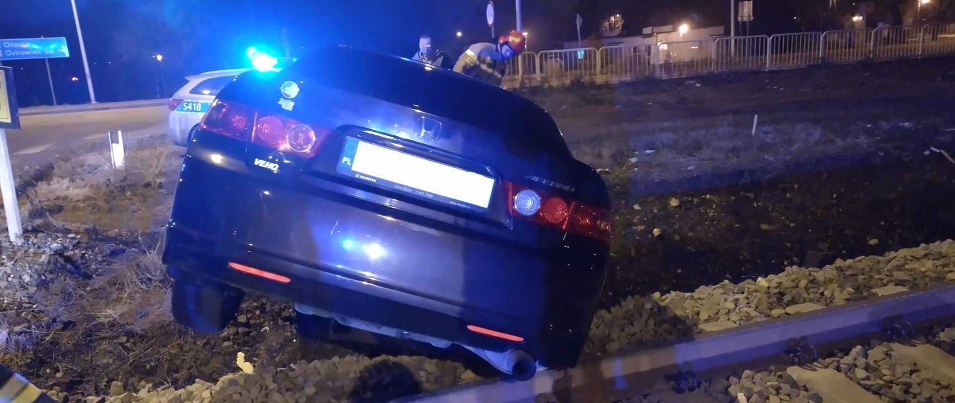 Samochód osobowy zawieszony tylną osia na torach kolejowych przy samochodzie z przodu znajduje się strażak. Pora nocna.