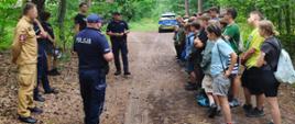 Grupa harcerzy stoi na leśnej drodze i słucha pogadanki prowadzonej przez funkcjonariuszy policji oraz przedstawicieli innych służb mundurowych. W tle radiowozy i drzewa otaczające miejsce spotkania.