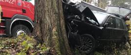 Zdjęcie przedstawia rozbity samochód osobowy marki BMW, który uderzył w przydrożne drzewo na tle samochodu pożarniczego.