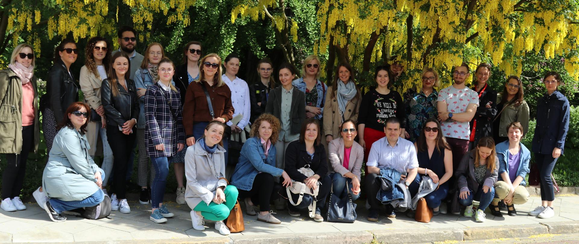  Zielone drzewa i kwitnące na żółto kwiaty w tle. Na środku stoi grupa kobiet i mężczyzn pozuje do zdjęcia. 